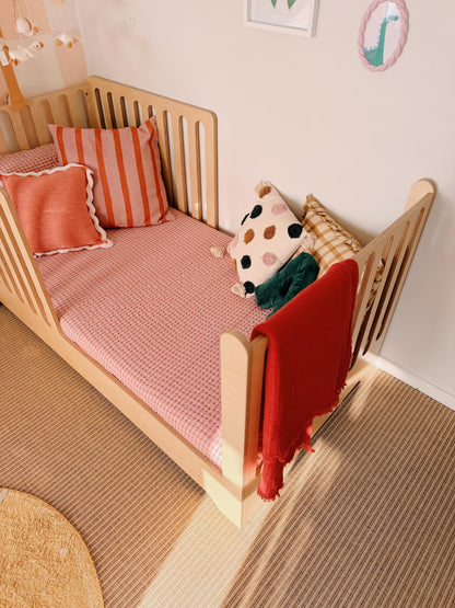 Children's bed with pink bedding, red blanket, and colorful pillows in a room.